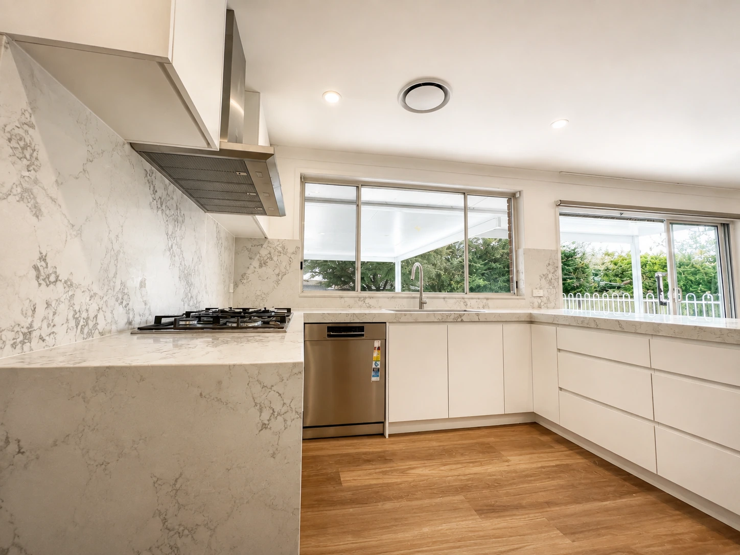 The same Castle Hill kitchen from the cooktop end, looking out to the pool — marble-look stone benchtops and a splashback carried to the ceiling, a wide canopy rangehood set into the bulkhead and white base cabinetry