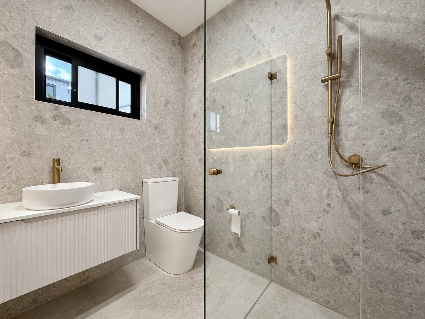 Second bathroom in the Strathfield home — terrazzo-look tiling, a frameless glass shower with brass fittings, a fluted wall-hung vanity and a black-framed slot window