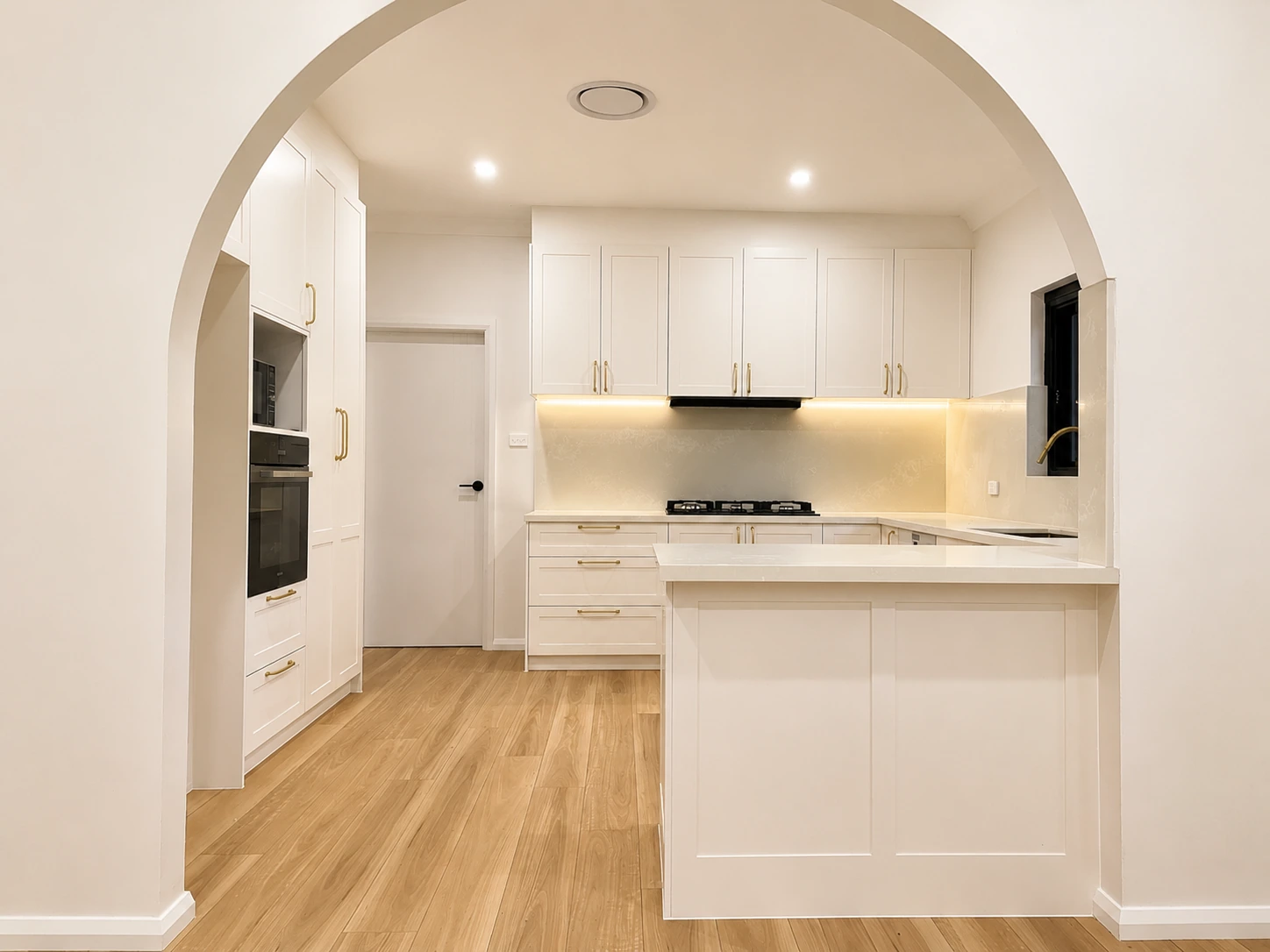 Whole-home renovation in Strathfield — looking through the retained archway into the new kitchen, with shaker overhead cupboards on brass handles, an integrated black rangehood, an LED-lit stone splashback and a wall oven and microwave tower