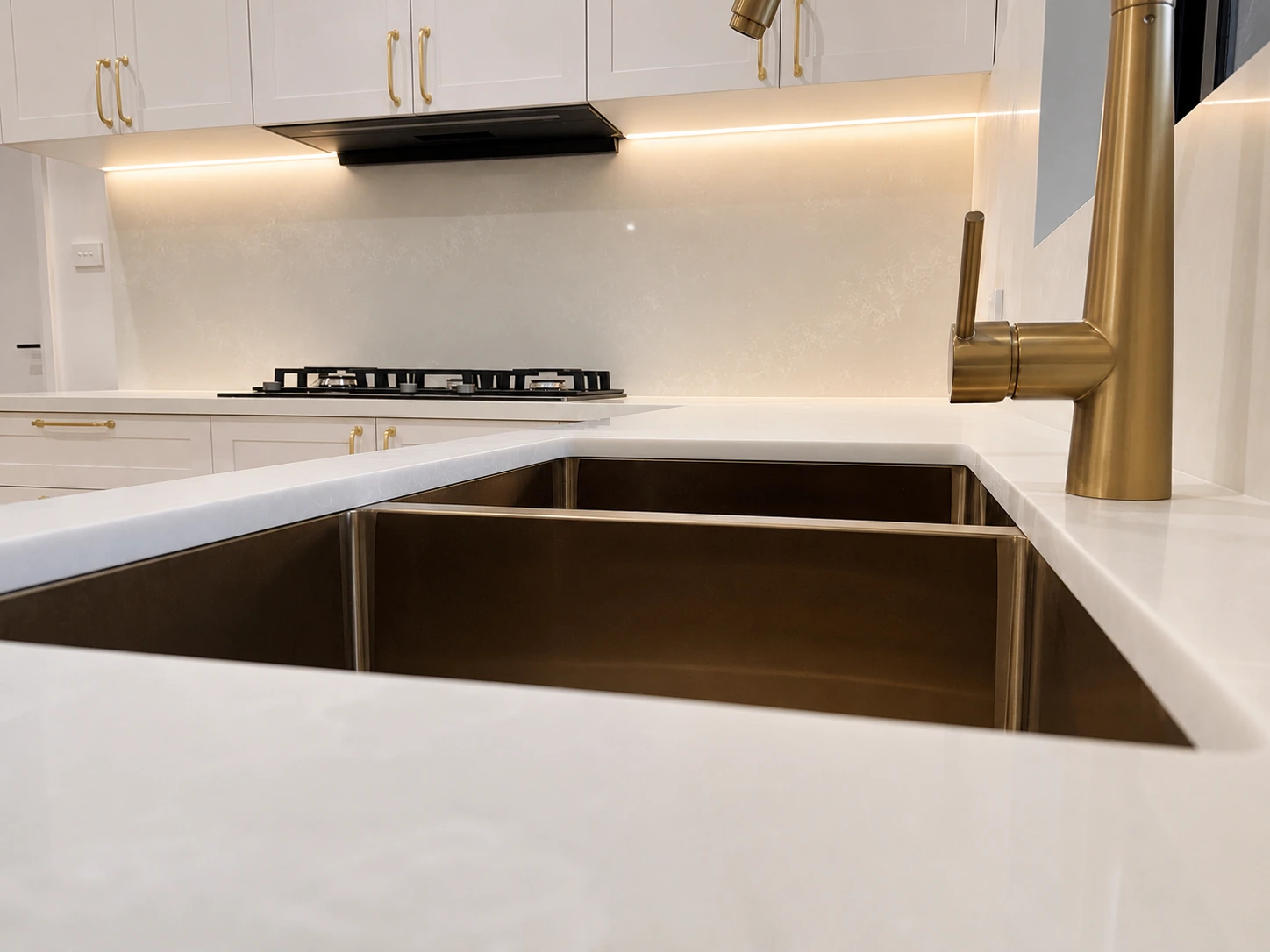 Close view of the Strathfield kitchen bench — a brushed brass gooseneck mixer over a brass undermount sink, with an LED strip lighting the stone splashback behind the cooktop