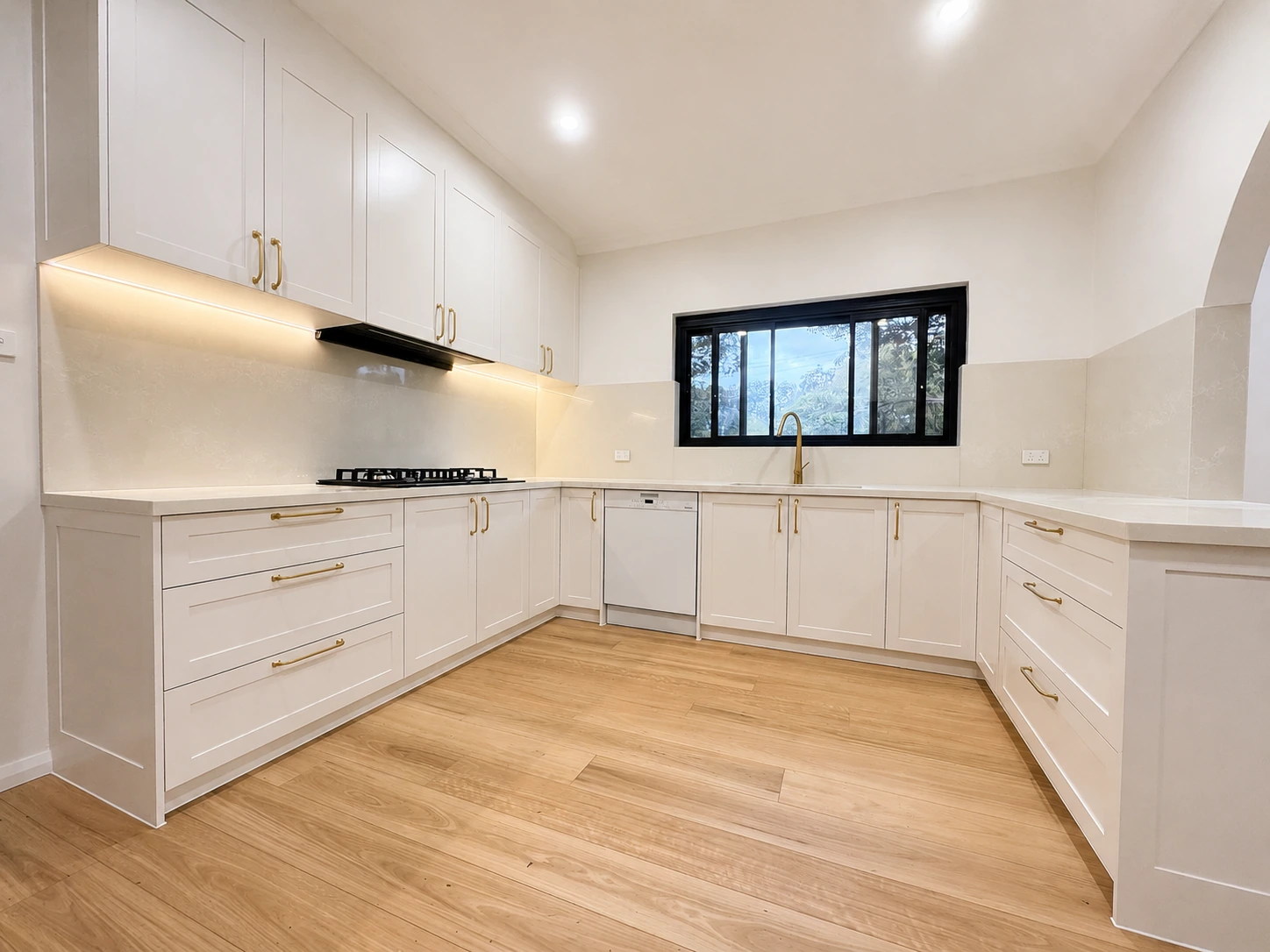 The Strathfield kitchen in full — U-shaped shaker joinery with brushed brass handles, stone benchtops and splashback, a gas cooktop and a black-framed window over the sink