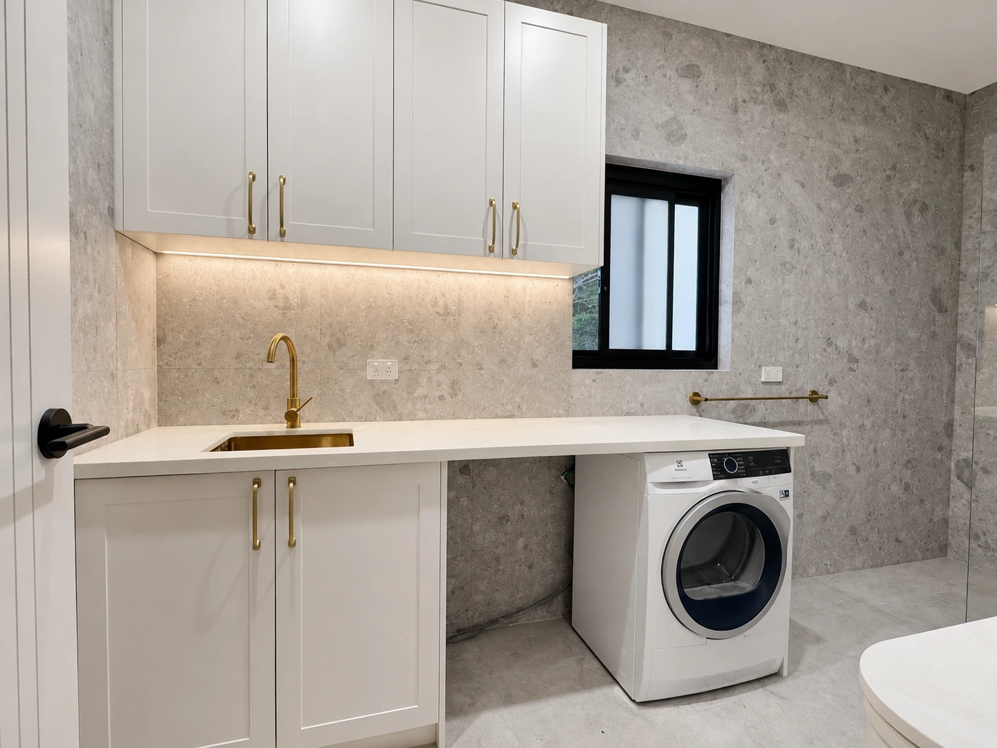 New laundry in the Strathfield renovation — shaker overhead cupboards, a stone benchtop with brass sink and mixer, terrazzo-look tiled walls and an under-bench washer