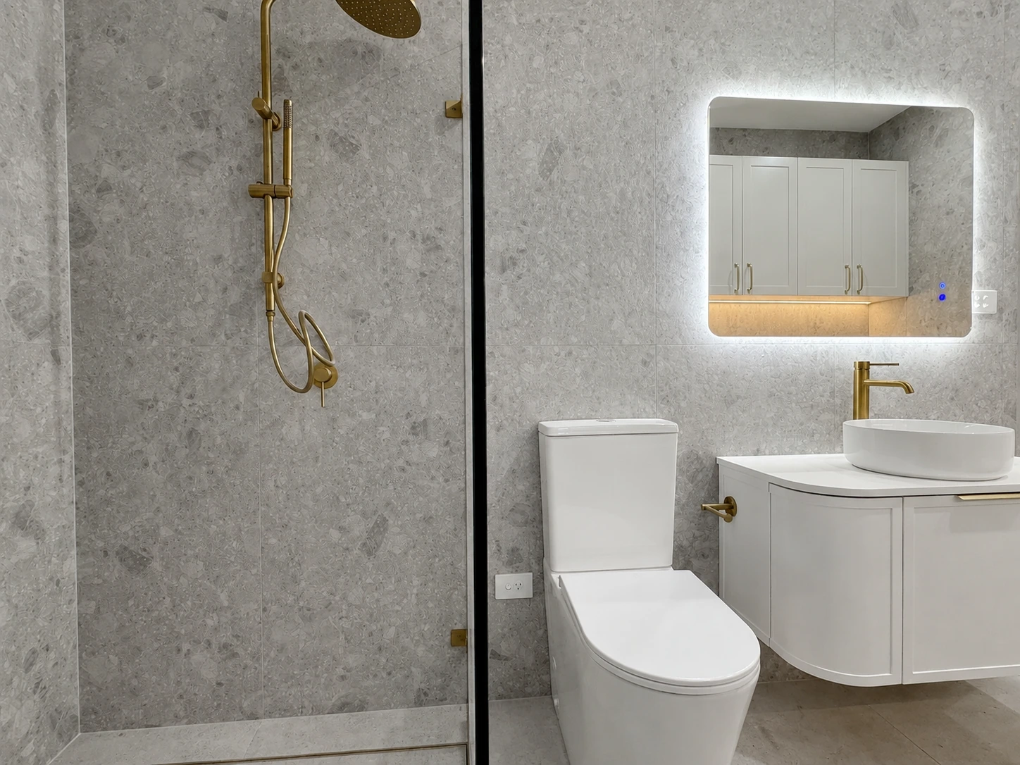Main bathroom in the Strathfield renovation — grey terrazzo-look tiles floor to ceiling, a brushed brass shower set, a backlit mirror and a curved wall-hung vanity with a vessel basin