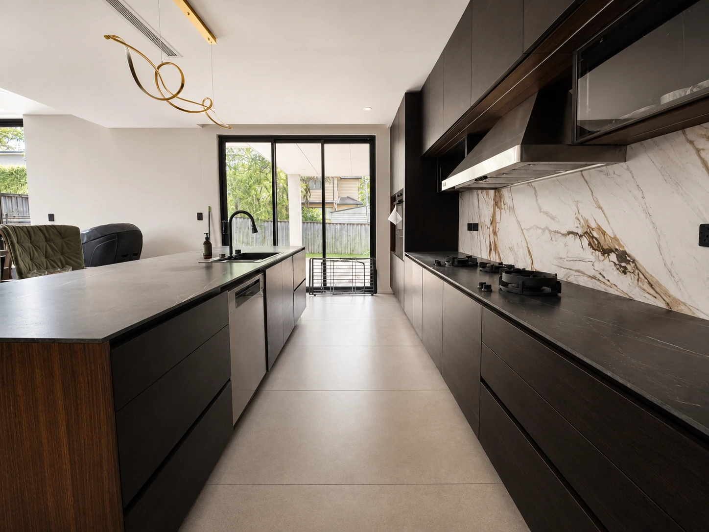 North Ryde galley kitchen — dark timber joinery, gold-veined marble splashback and a sculptural brass pendant opening to the garden