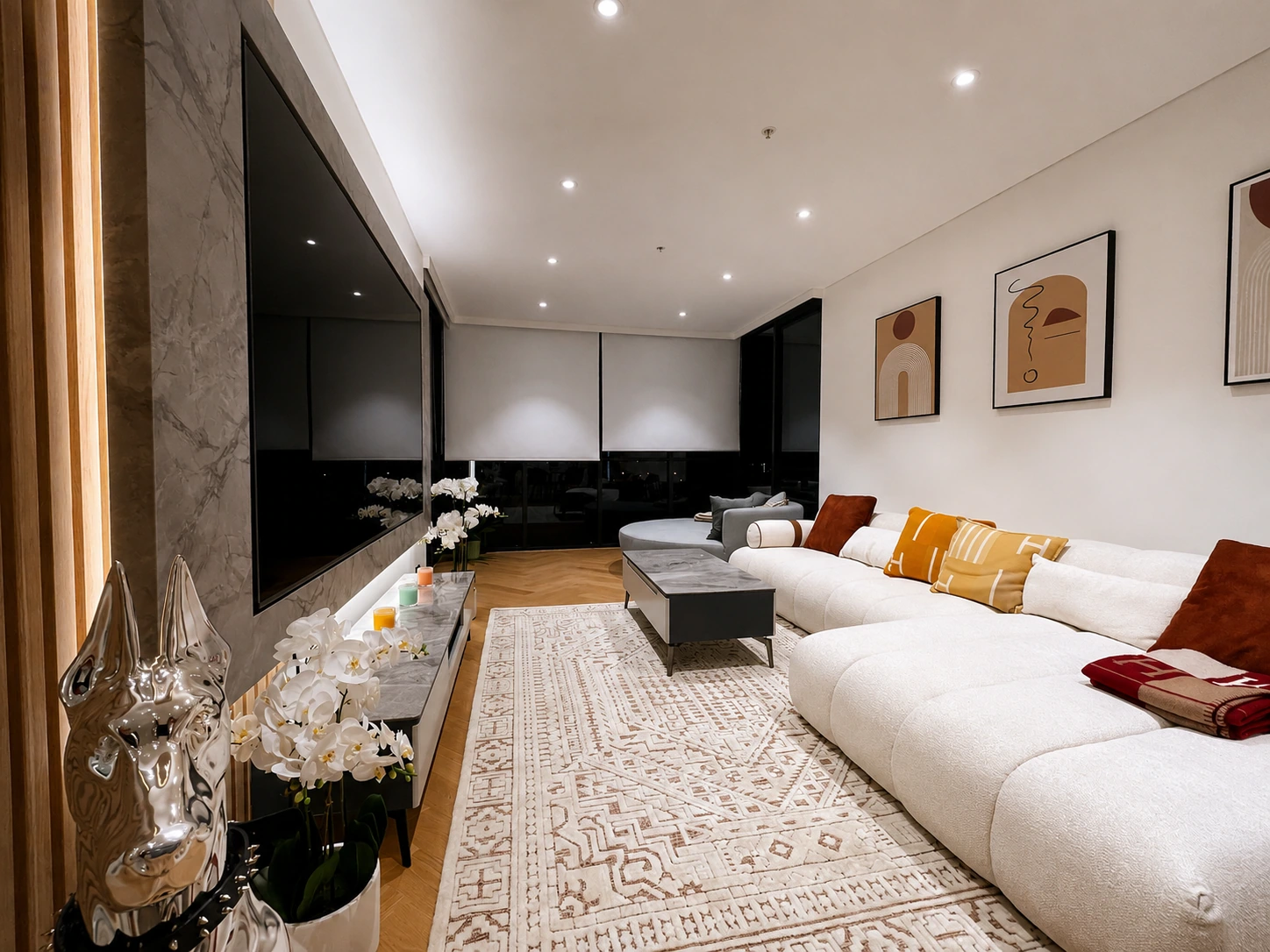 Wide view down the length of the Rhodes apartment living room at night — herringbone oak floors, the stone television feature wall with concealed lighting on the left, and a cream boucle modular sofa running to the windows