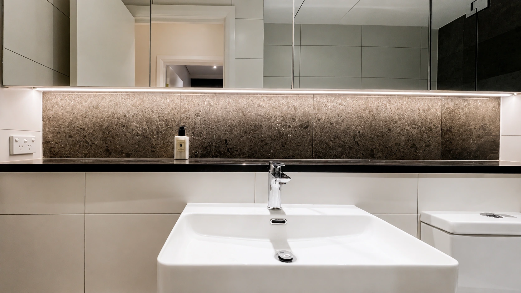 Rhodes apartment bathroom — mirrored shaving cabinets with an LED strip washing the fossil-stone splashback, over a black stone benchtop and a square white basin