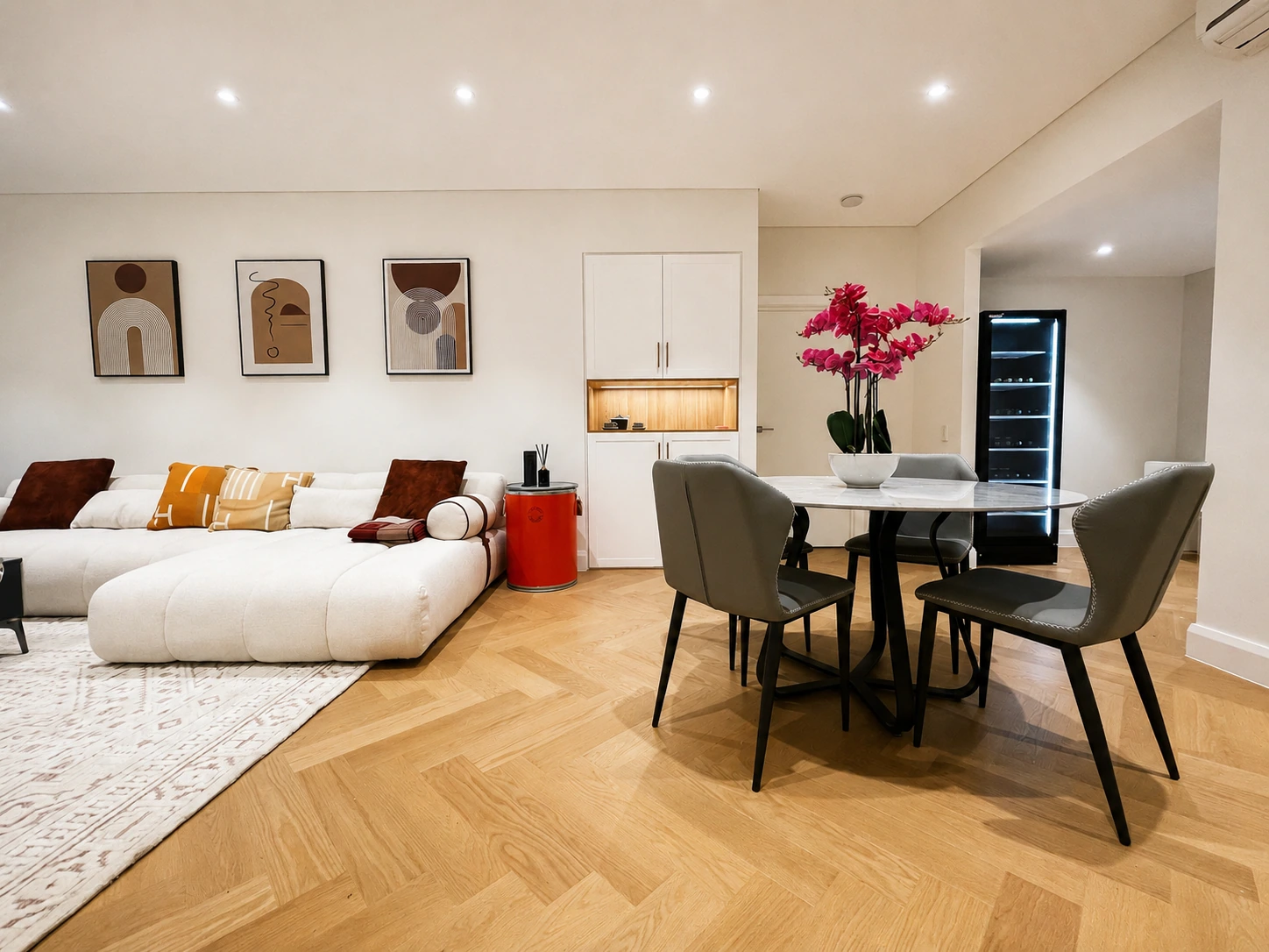 Apartment renovation in Rhodes — daylit open living and dining with herringbone oak floors, custom white joinery holding a lit oak niche, and a round marble table beside a freestanding glass-door wine cabinet