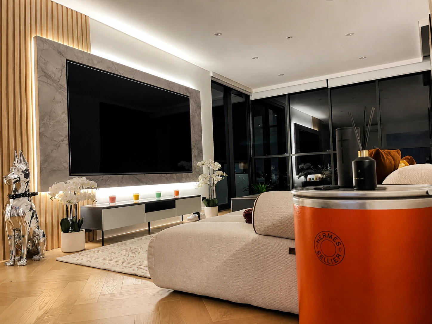 Corner view of the Rhodes apartment living room at night — the lit stone television wall and fluted oak battens beside the dark full-height glazing, with an orange drum side table in the foreground